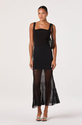 A woman is wearing a sleeveless black top with wide shoulder straps and a square neckline. She pairs it with a fitted black mini skirt that extends just above the knees. She is also wearing sheer, black, vertically striped pants that reach her ankles, creating a layered look. The outfit is complemented by white open-toe sandals with black embellishments.